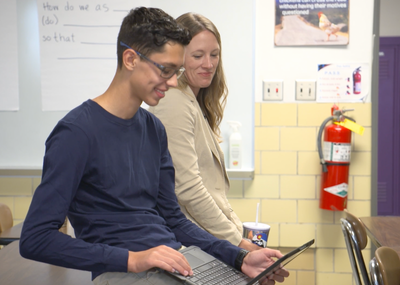 Photograph of two educators in a classroom, one smiling at their laptop computer.